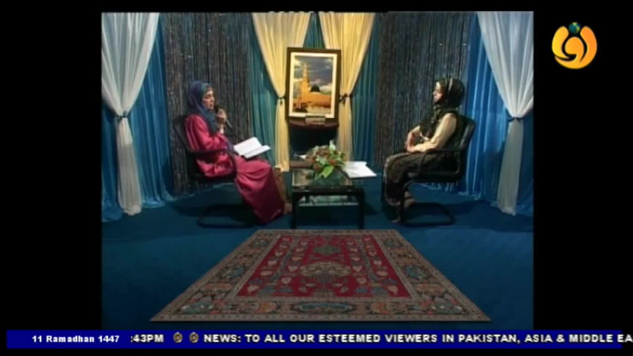 Two women sit on chairs facing each other across a coffee table. A microphone is held by the woman on the left, who is looking at a book. Two women sit on chairs facing each other across a coffee table. A microphone is held by the woman on the left, who is looking at a book.