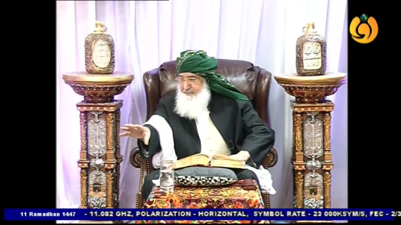 A man in a green turban gestures with his left hand while speaking. He sits in a leather chair with a book open before him, a glass of water to his side.