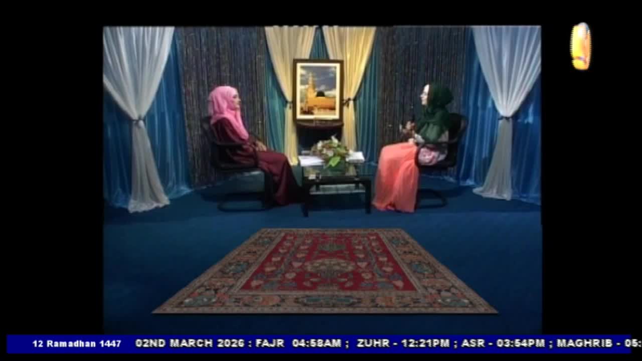 Two women sit opposite each other on a blue carpeted set, a framed picture of a mosque behind them. One woman wears a pink hijab and a maroon dress, while the other has a green hijab and a peach-colored skirt.