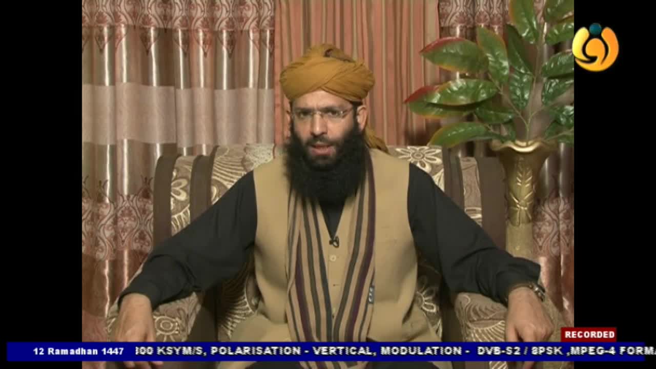 A man with a beard and glasses, wearing a turban and vest, speaks directly to the camera. He is seated in front of striped curtains, with a potted plant to his right.