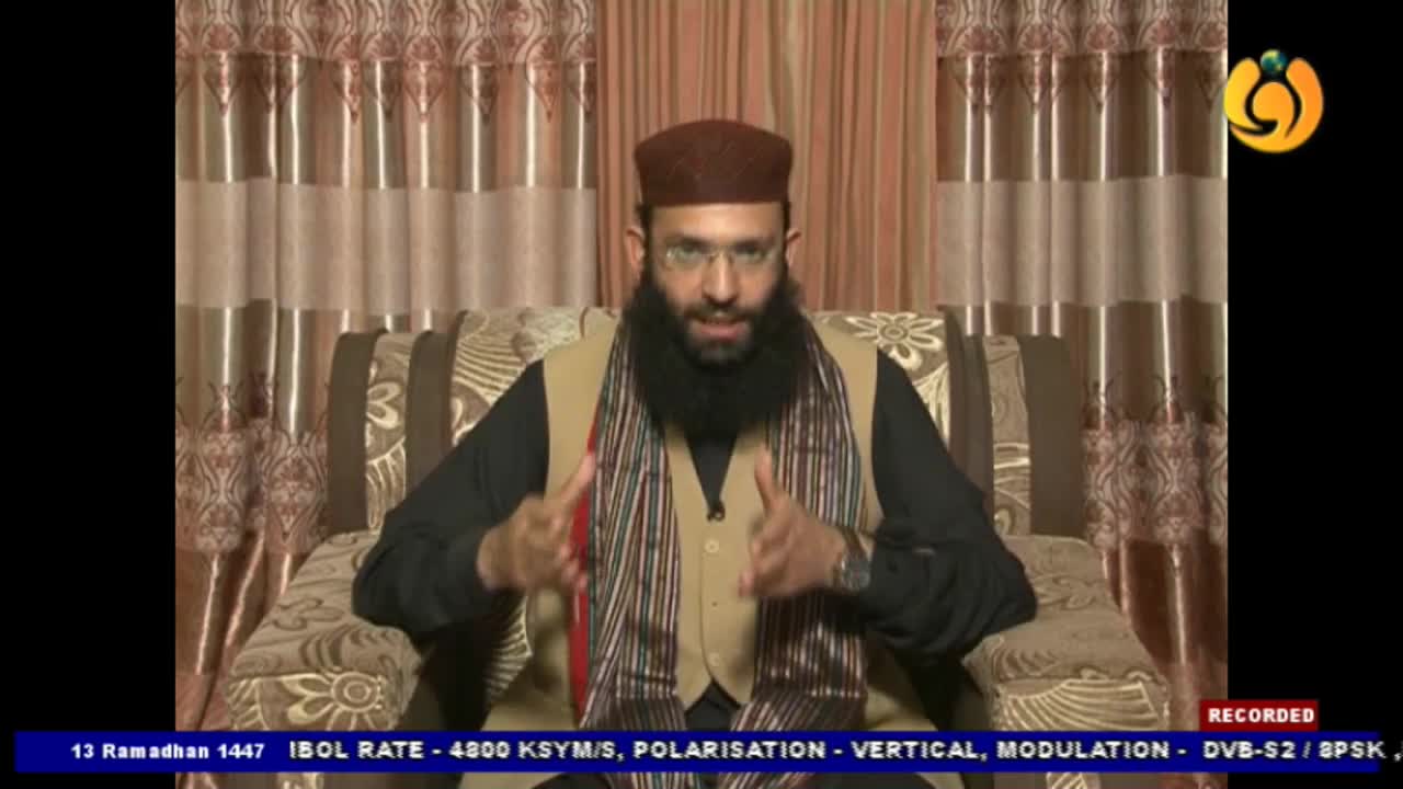 A man with a beard and glasses, wearing a brown cap and a striped scarf, gestures with his hands. He's seated in a patterned armchair, with curtains behind him.