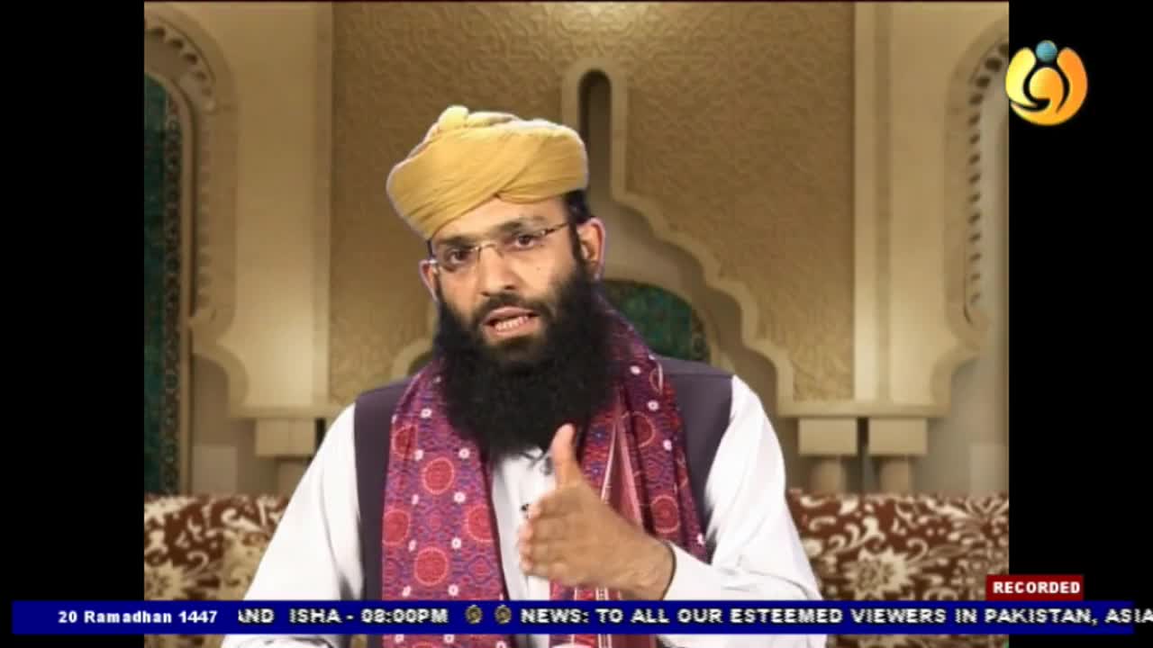 A man with a beard and glasses, wearing a yellow turban and a patterned scarf, gestures with his right hand. He speaks directly to the camera, likely for a Noor TV broadcast.