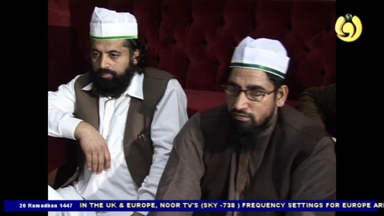 Two men in white kufi hats sit close together, one with a dark beard and the other with glasses and a beard. Their expressions are serious as they look off to the side, their shoulders slightly slumped.