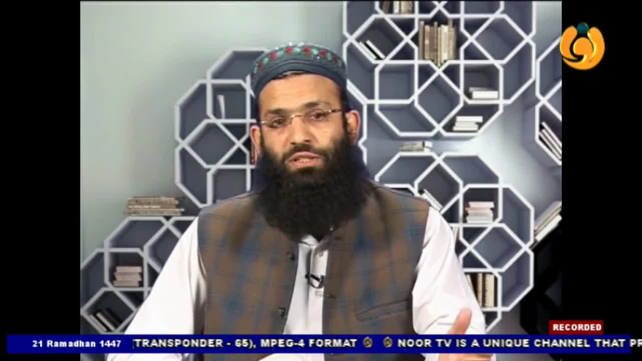 A man with a beard and glasses, wearing a skullcap and vest, speaks directly to the camera. Behind him, a geometric bookshelf displays several books.
