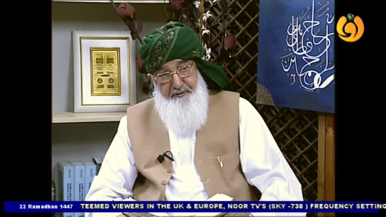 An elder with a long white beard and a green turban speaks, wearing a tan waistcoat over a white garment. He sits in front of a framed gold plaque and a blue canvas with white Arabic calligraphy.