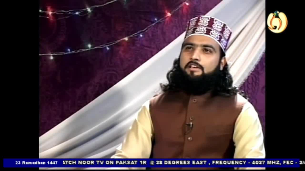 A man with a long beard and a patterned cap speaks on Noor TV. He wears a brown vest over a cream-colored shirt.