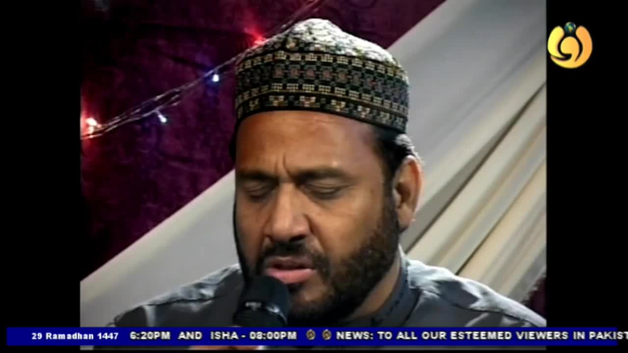 A man wearing a patterned cap speaks into a microphone, his eyes closed in concentration. Behind him, small, colorful lights twinkle against a dark fabric. A man wearing a patterned cap speaks into a microphone, his eyes closed in concentration. Behind him, small, colorful lights twinkle against a dark fabric.