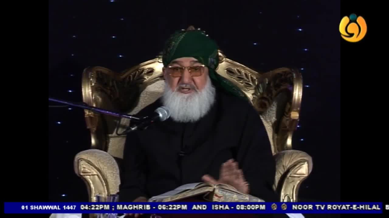 A man with a long white beard, wearing a green turban and glasses, speaks into a microphone while seated on an ornate chair. He gestures with his right hand, his gaze directed forward, with an open book before him. A man with a long white beard, wearing a green turban and glasses, speaks into a microphone while seated on an ornate chair. He gestures with his right hand, his gaze directed forward, with an open book before him.