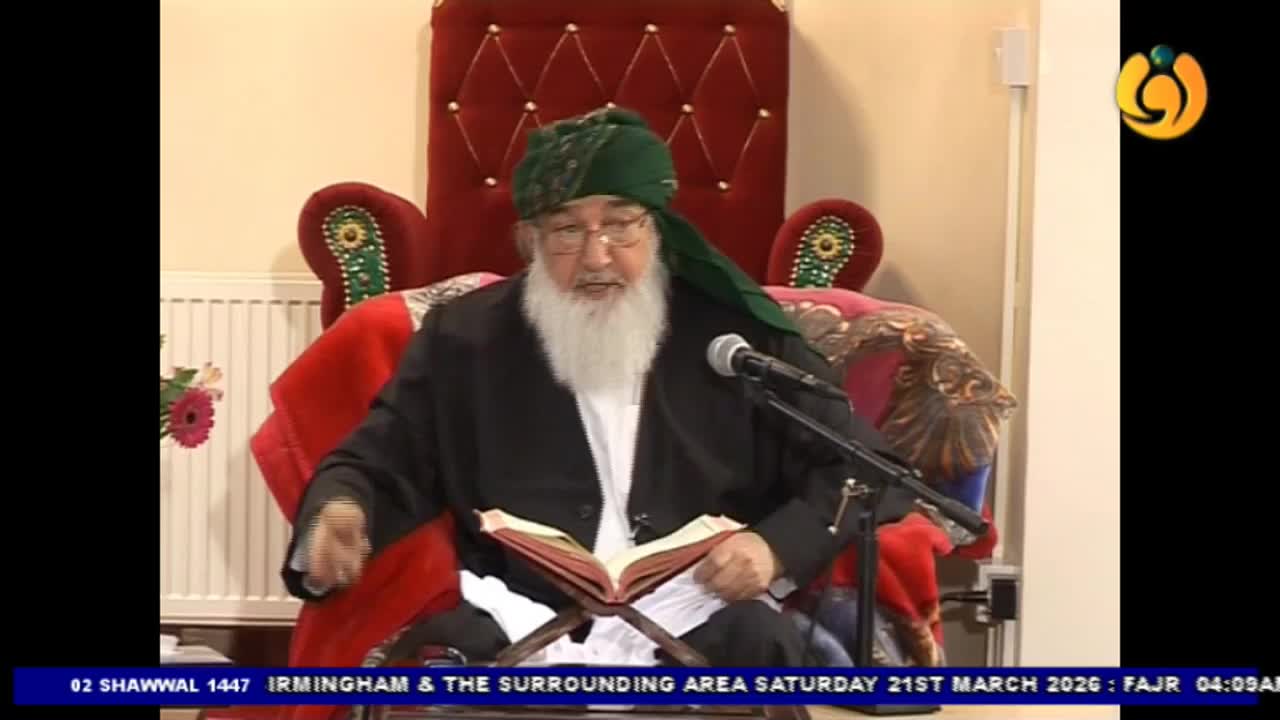 An older man wearing a green turban and glasses speaks into a microphone, his hands resting on an open book before him. He sits in a plush, red chair, a scene broadcast by Noor TV from Irirmingham. An older man wearing a green turban and glasses speaks into a microphone, his hands resting on an open book before him. He sits in a plush, red chair, a scene broadcast by Noor TV from Irirmingham.