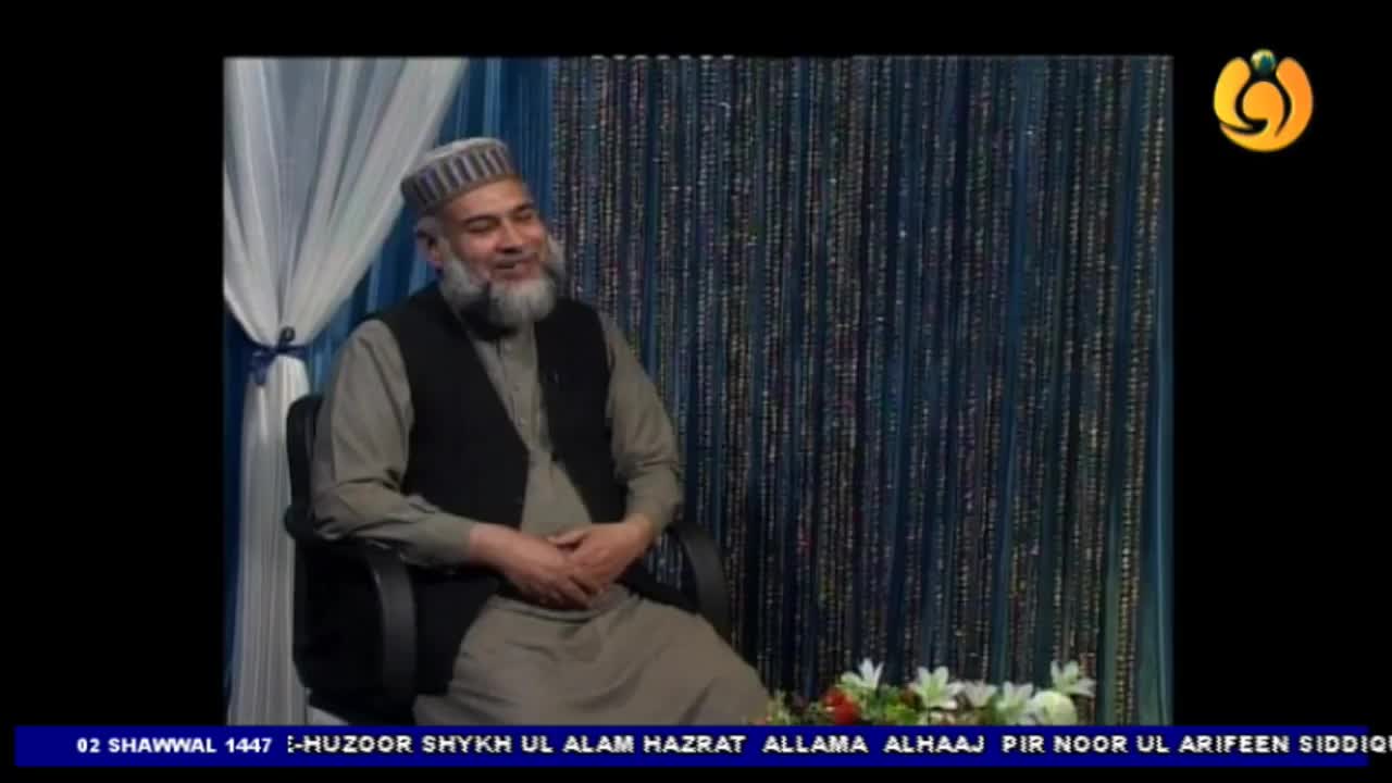 A man with a grey beard and a patterned cap sits in a chair, speaking. He wears a grey tunic and a dark vest, his hands resting on his lap. The background behind him is a shimmering curtain of blue and silver strands. A man with a grey beard and a patterned cap sits in a chair, speaking. He wears a grey tunic and a dark vest, his hands resting on his lap. The background behind him is a shimmering curtain of blue and silver strands.
