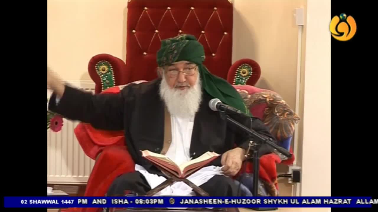 An elderly man in a green turban gestures with his left hand while speaking into a microphone. He sits on a red chair, a book open on a stand before him. An elderly man in a green turban gestures with his left hand while speaking into a microphone. He sits on a red chair, a book open on a stand before him.