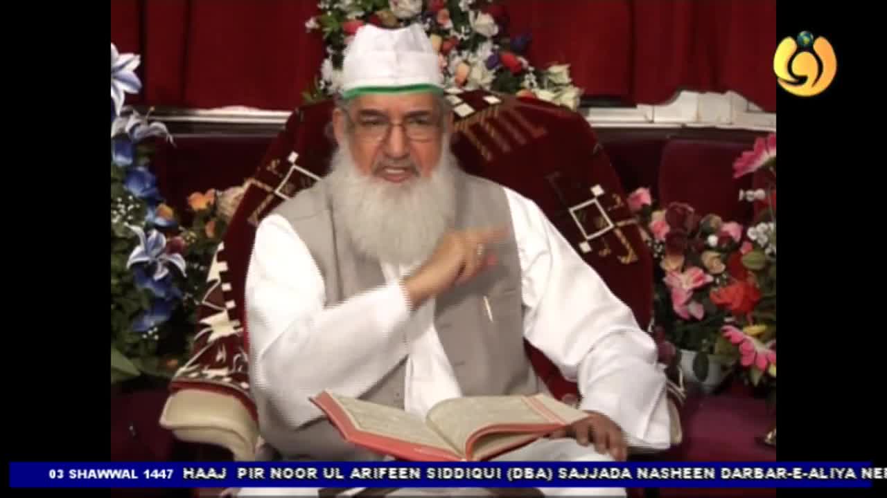 A man with a long white beard, wearing a white cap and vest over a white shirt, gestures with his right hand while speaking. He sits in a plush chair with an open book before him, likely during a broadcast for Noor TV. A man with a long white beard, wearing a white cap and vest over a white shirt, gestures with his right hand while speaking. He sits in a plush chair with an open book before him, likely during a broadcast for Noor TV.