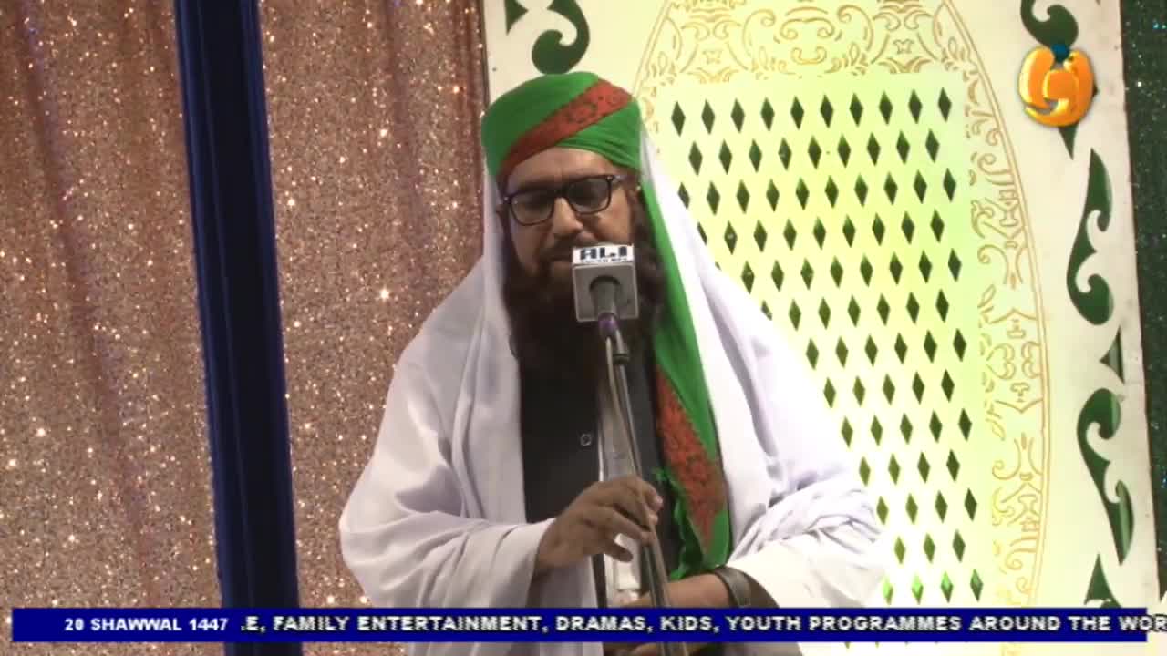 A man in a green turban and white robe stands at a microphone. He is speaking into the mic, his face partially obscured by his beard. A man in a green turban and white robe stands at a microphone. He is speaking into the mic, his face partially obscured by his beard.