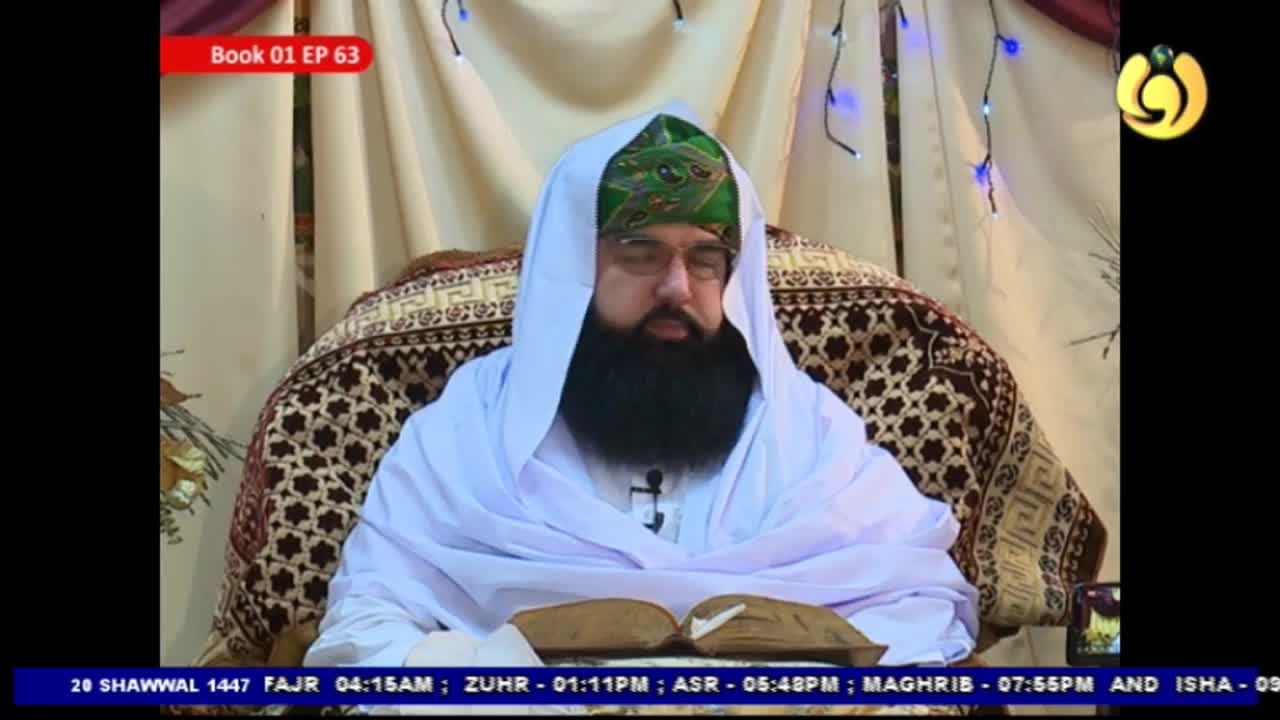 A man with a long black beard and glasses sits, draped in white fabric and wearing a green turban. He's looking down at an open book in front of him, with a patterned cloth behind him. A man with a long black beard and glasses sits, draped in white fabric and wearing a green turban. He's looking down at an open book in front of him, with a patterned cloth behind him.