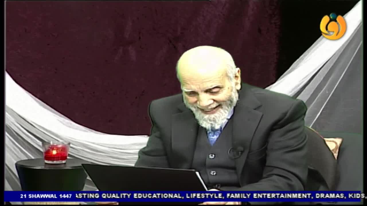 A man with a white beard and bald head, dressed in a suit, laughs heartily while looking at a laptop. A lit red candle sits on a small table to his left, and a white draped fabric hangs behind him, suggesting a studio setting for Noor TV. A man with a white beard and bald head, dressed in a suit, laughs heartily while looking at a laptop. A lit red candle sits on a small table to his left, and a white draped fabric hangs behind him, suggesting a studio setting for Noor TV.