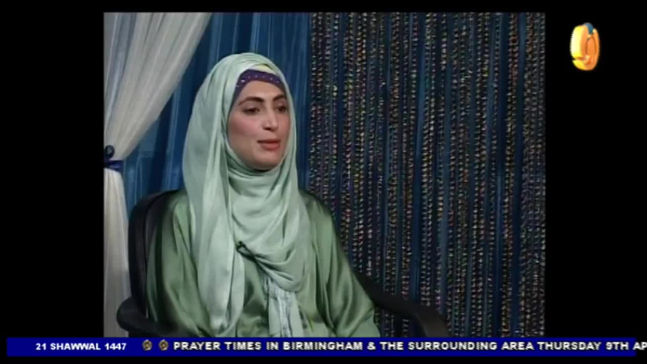 A woman wearing a light green hijab and dress speaks into a microphone. The background features a beaded curtain and a small gold logo in the corner. A woman wearing a light green hijab and dress speaks into a microphone. The background features a beaded curtain and a small gold logo in the corner.