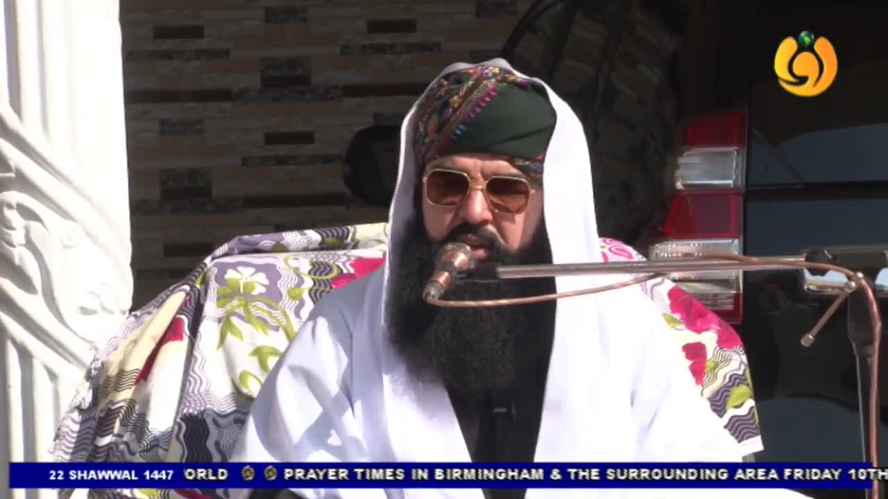 A man with a long beard and sunglasses speaks into a microphone, his head covered by a white cloth and a patterned turban. He is seated on a chair draped with a colourful fabric, with a black vehicle visible behind him. A man with a long beard and sunglasses speaks into a microphone, his head covered by a white cloth and a patterned turban. He is seated on a chair draped with a colourful fabric, with a black vehicle visible behind him.