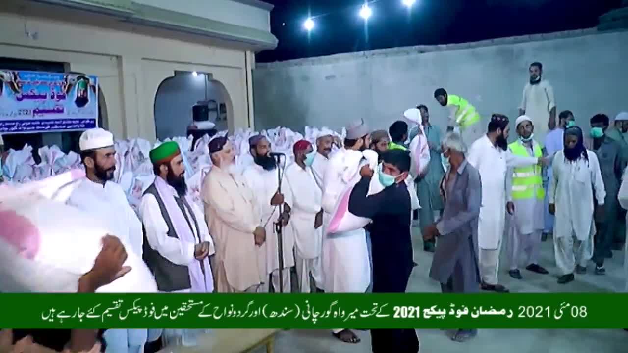 Men in traditional clothing are handing out large white bags of what looks like food. A banner in Urdu hangs in the background, announcing a food distribution for Ramadan 2021. Men in traditional clothing are handing out large white bags of what looks like food. A banner in Urdu hangs in the background, announcing a food distribution for Ramadan 2021.