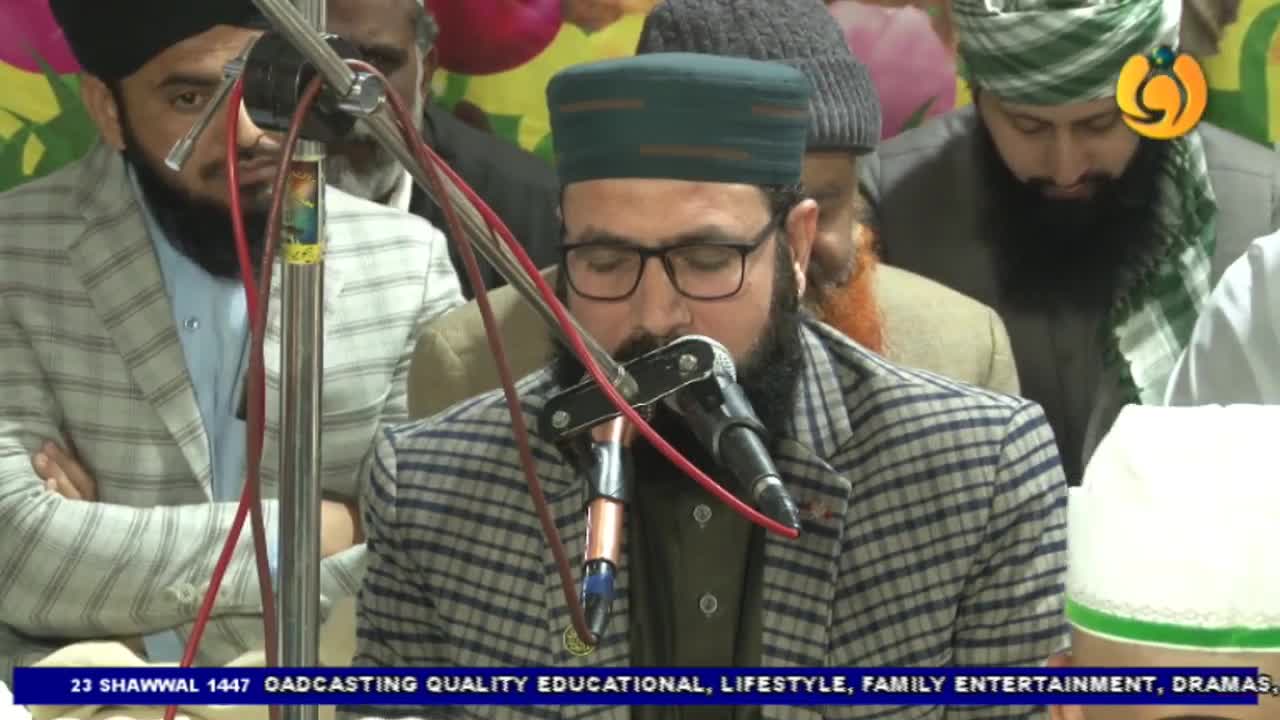 A man in a checkered jacket and cap reads from a book, his voice amplified by a microphone. The Noor TV logo is visible in the corner, indicating a broadcast from the UK. A man in a checkered jacket and cap reads from a book, his voice amplified by a microphone. The Noor TV logo is visible in the corner, indicating a broadcast from the UK.