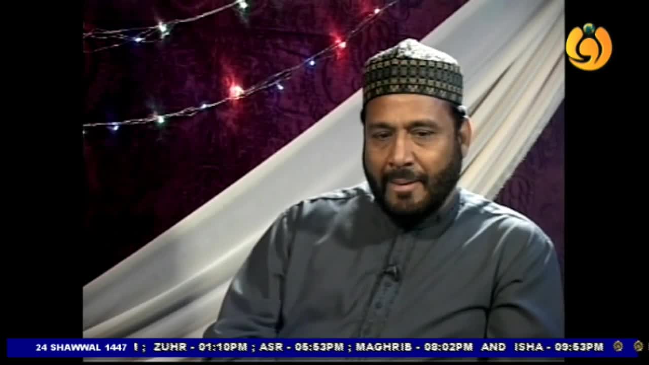 A man with a beard and a patterned cap is speaking. String lights with small bulbs are draped behind him.