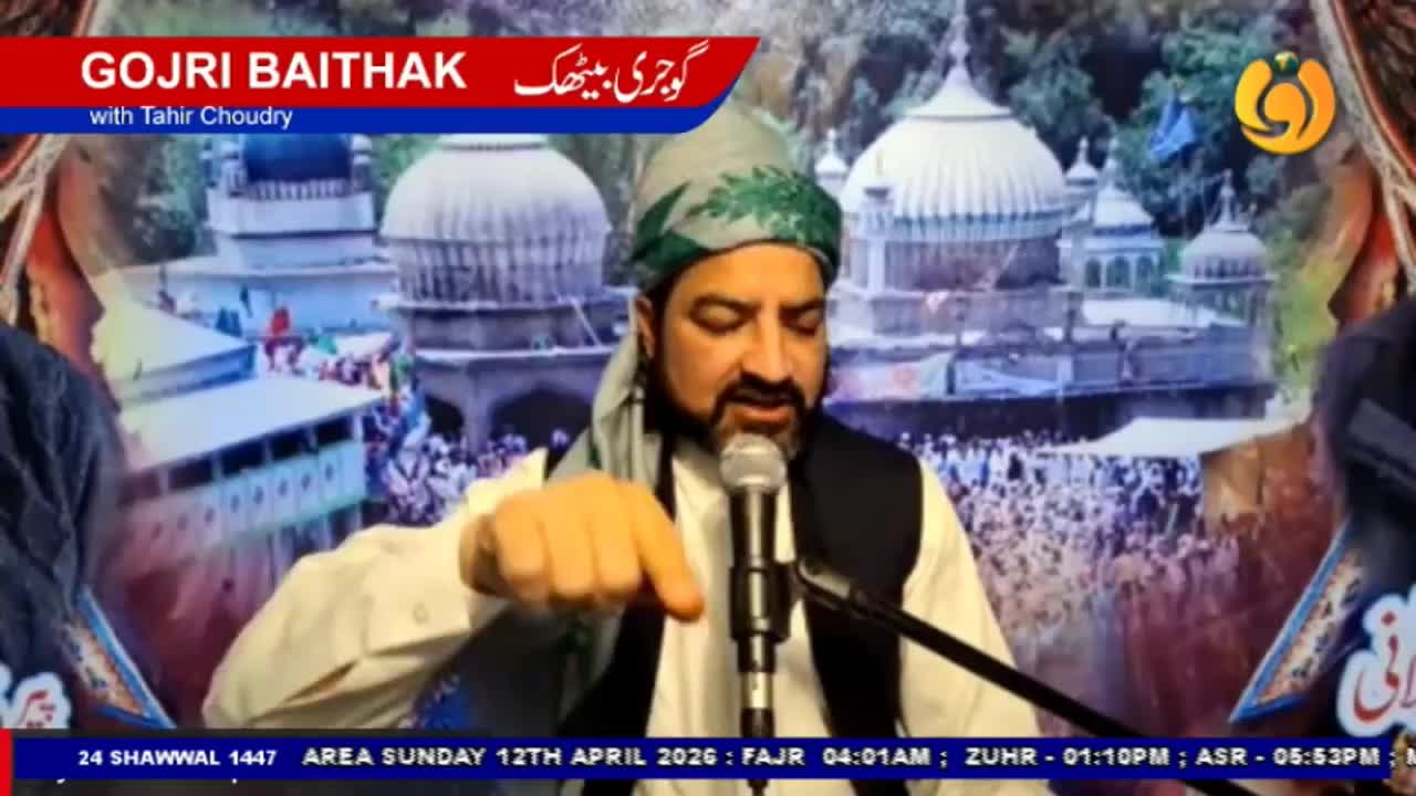A man in a green turban speaks into a microphone, gesturing with his right hand. Behind him, a crowd gathers before ornate domed buildings, a scene broadcast on Noor TV.