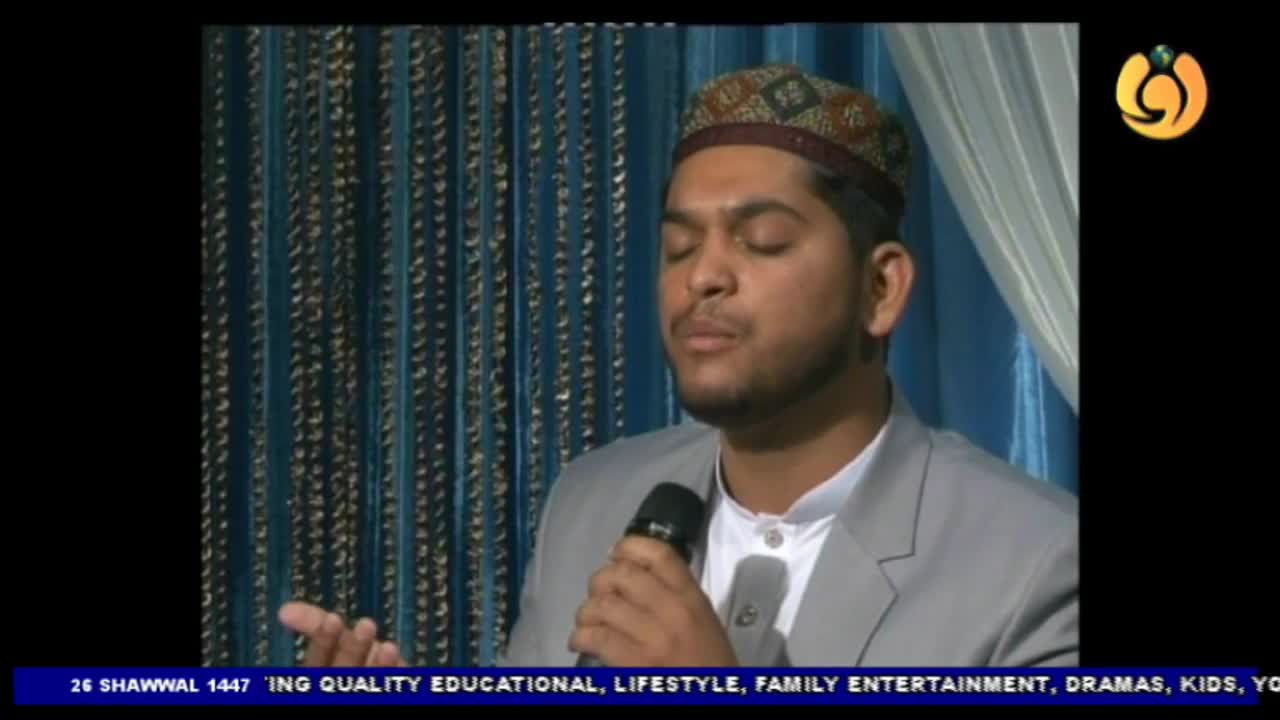 A man in a patterned cap and grey suit holds a microphone, his eyes closed as he speaks. The Noor TV logo appears in the corner, with text at the bottom indicating the date and programming.