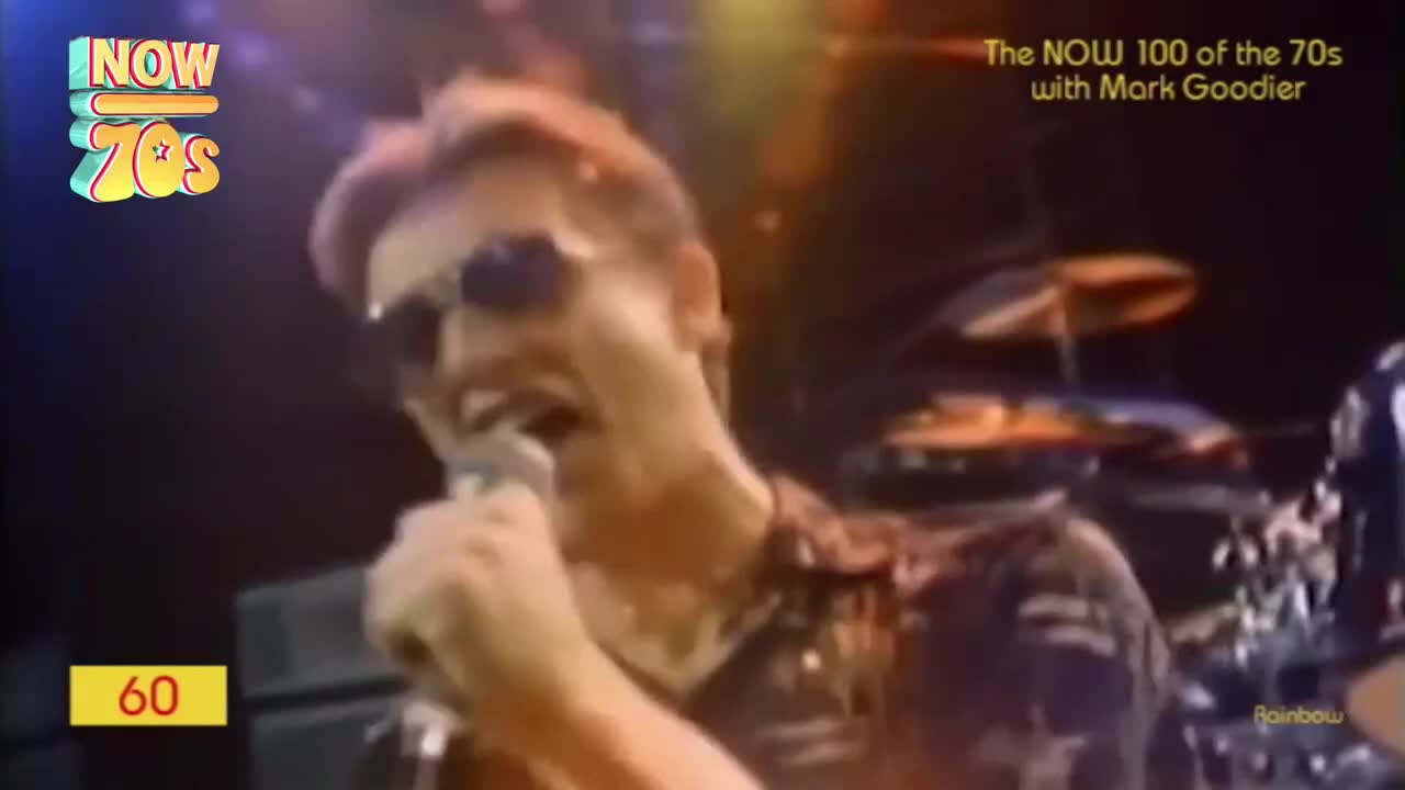 The singer, sporting sunglasses and a patterned shirt, leans into the microphone, belting out a tune. Behind him, the drummer is visible, poised to keep the rhythm going.
