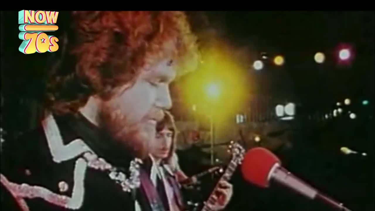 The singer, with his red hair and beard, is leaning into the microphone, while a guitarist strums behind him. Bright stage lights illuminate the band as they perform.
