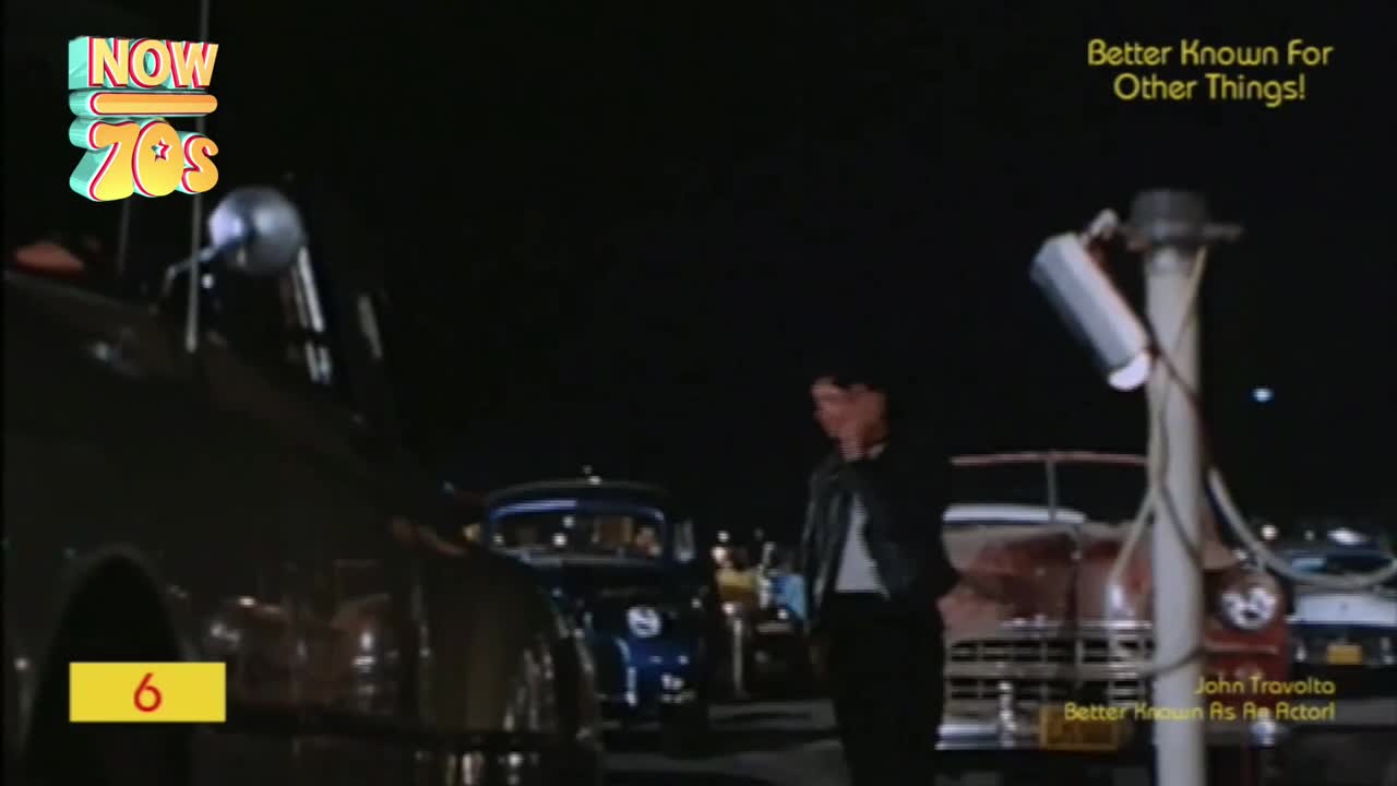 A man in a leather jacket stands near a line of cars, illuminated by harsh overhead lights. The dark night provides the backdrop for a scene that feels like it's straight out of the 70s.

