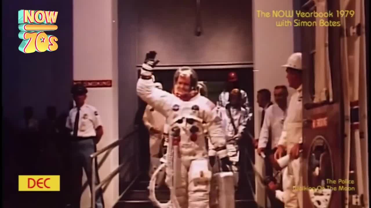 A man in a bulky spacesuit waves as he emerges from a doorway, surrounded by people in white coats and uniforms. The scene is being filmed for a TV show from the United Kingdom, back in the 70s.
