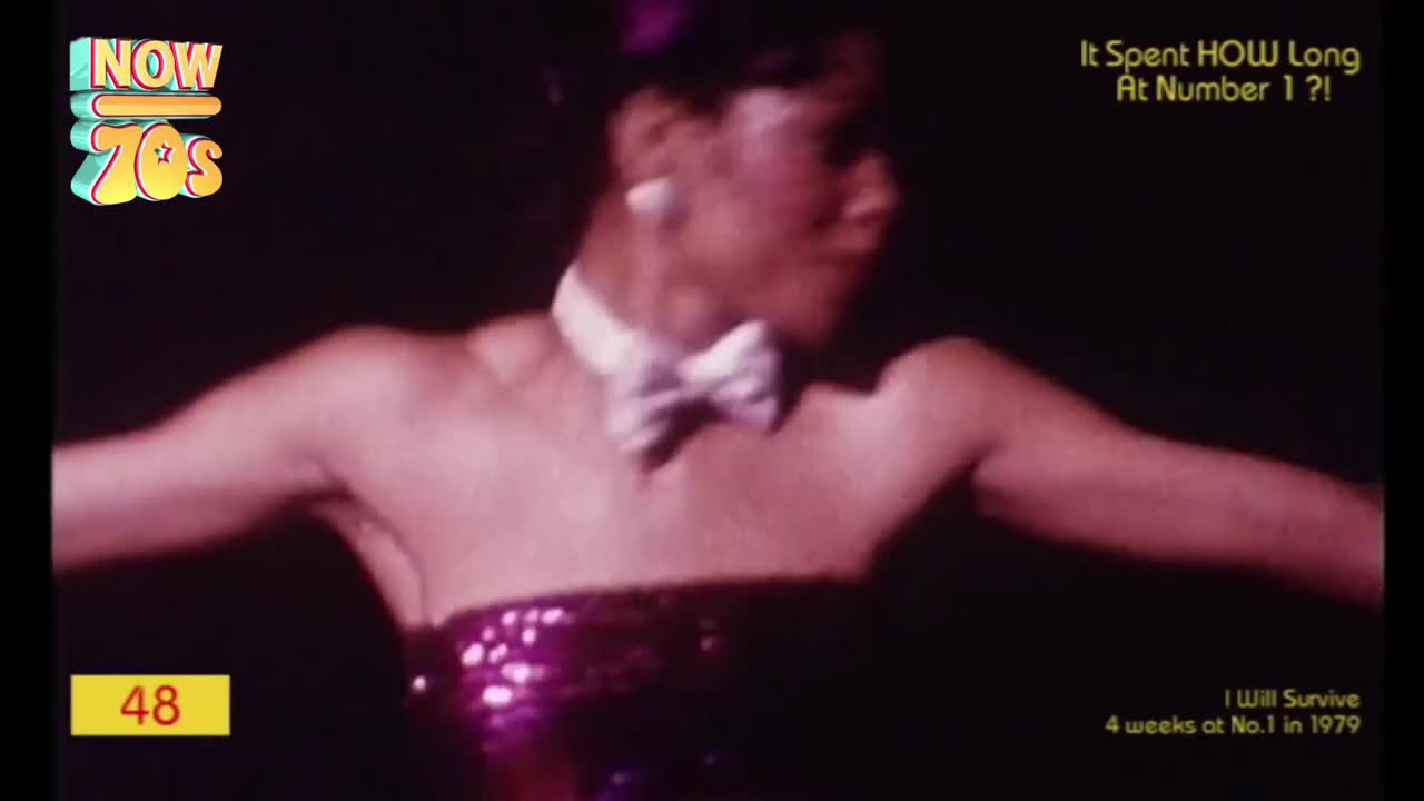 A woman, in a sparkly pink dress, is dancing, arms outstretched. She's wearing a bow tie, and the camera's zoomed in close.
