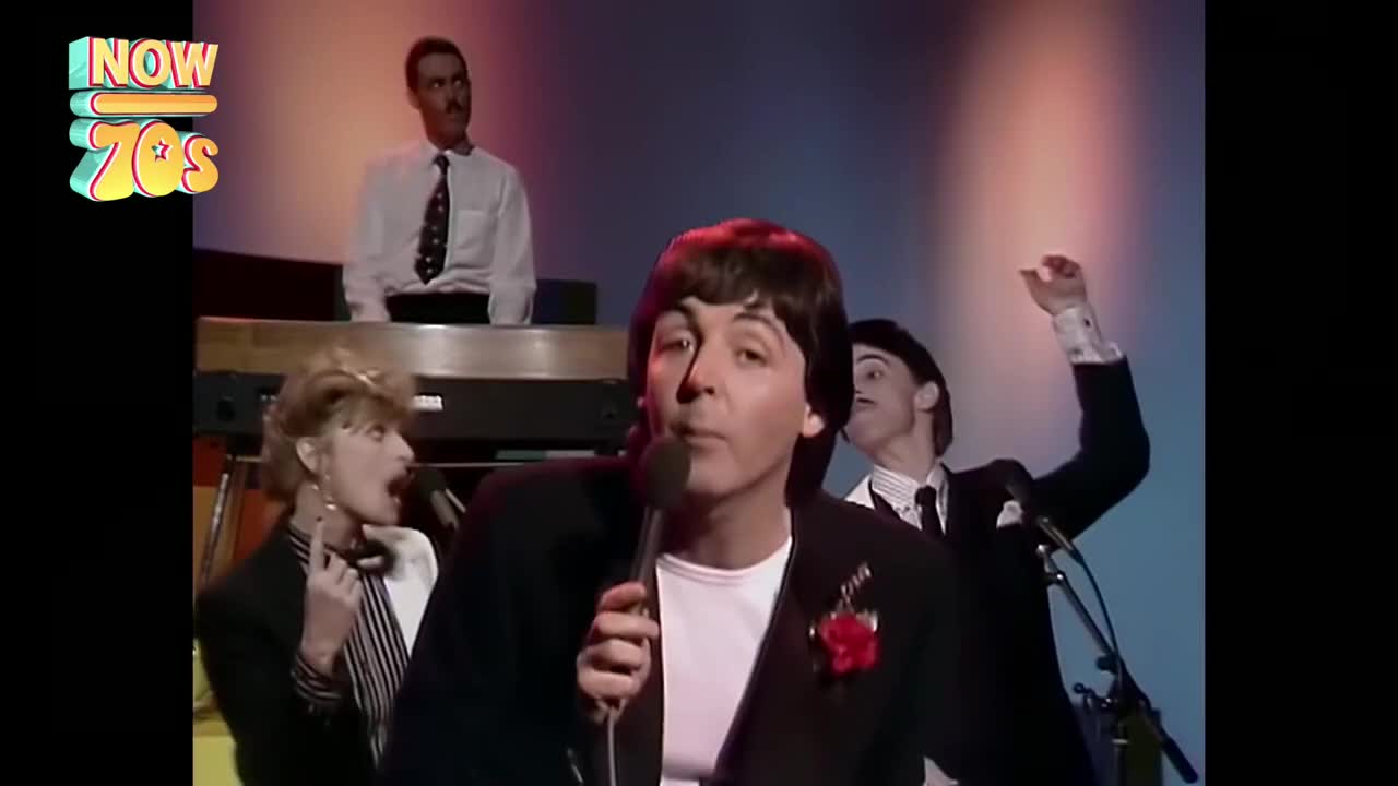 The singer, wearing a black suit with a red flower, is holding a microphone, while the band members around him are playing. A keyboard player is behind him, and the drummer raises his arm in the air.
