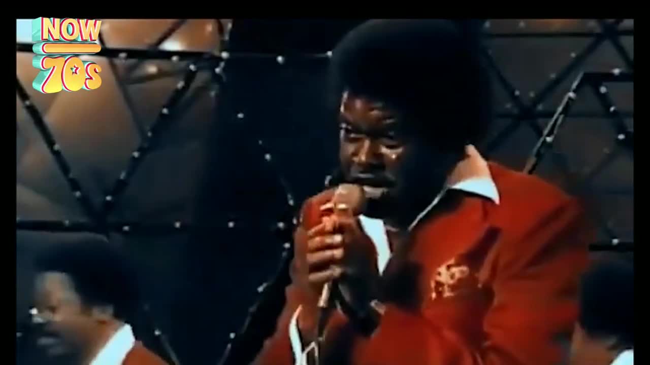 The singer, drenched in sweat, grips the microphone tightly, eyes closed as he belts out a tune. His red jacket and the band behind him are lit by the stage lights.
