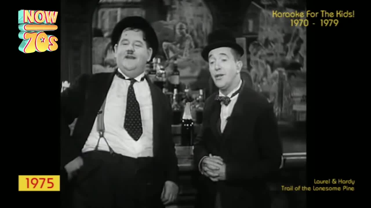 Two men in suits and bowler hats stand side-by-side, one with a polka-dot tie and the other with a bow tie. They're in a black-and-white scene from a film, likely from the United Kingdom, and it looks like it's from the 70s.
