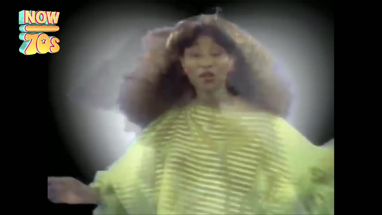 A woman in a striped, pale green top is singing, her hair billowing around her head. The background is a hazy, greyish-white, like a studio effect from the UK in the 70s.
