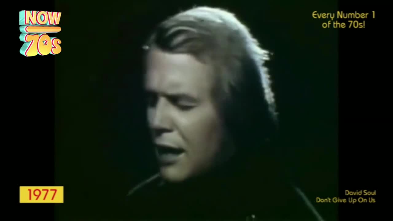 A man with long, wavy hair sings into a microphone, his face illuminated against a dark backdrop. The screen displays "Now 70s" and the year 1977.
