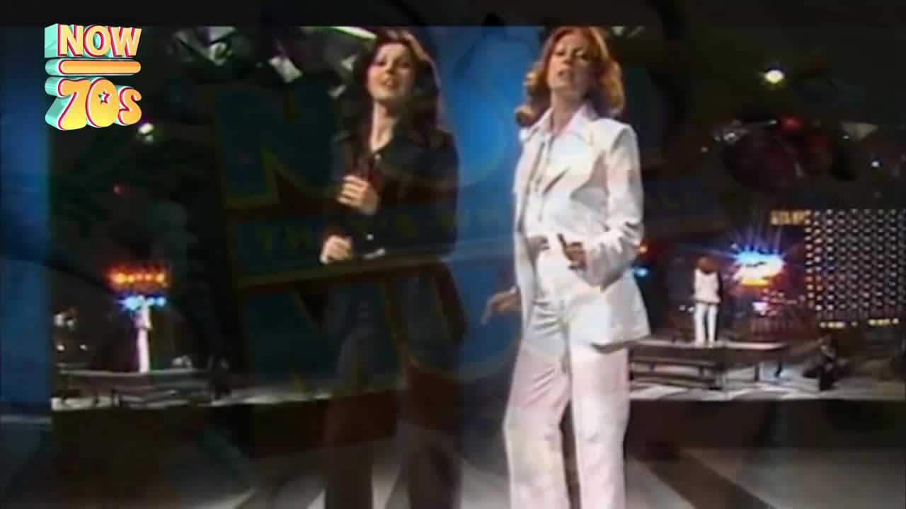 On a brightly lit stage, two women in sharp suits are singing, gesturing towards the audience. A smaller figure in white stands on a raised platform at the back, bathed in stage lights.
