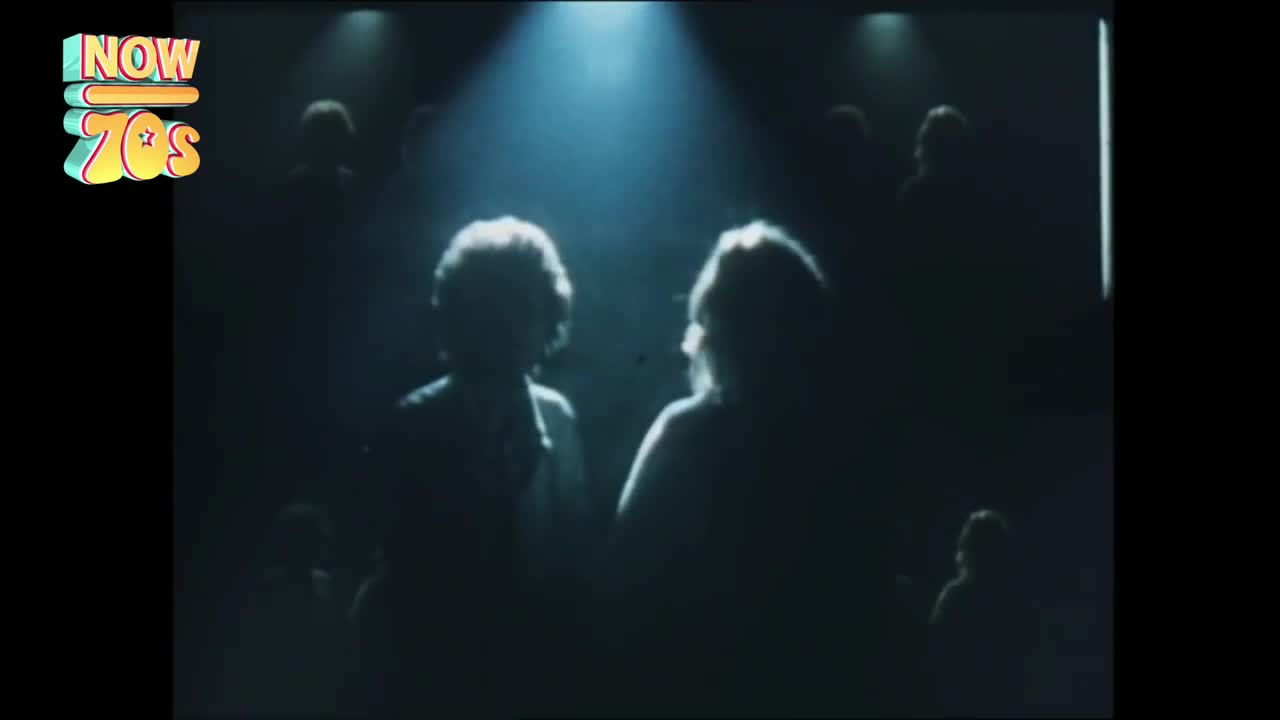 Two figures stand in a spotlight, their backs to the camera. The rest of the audience is barely visible in the dimness.
