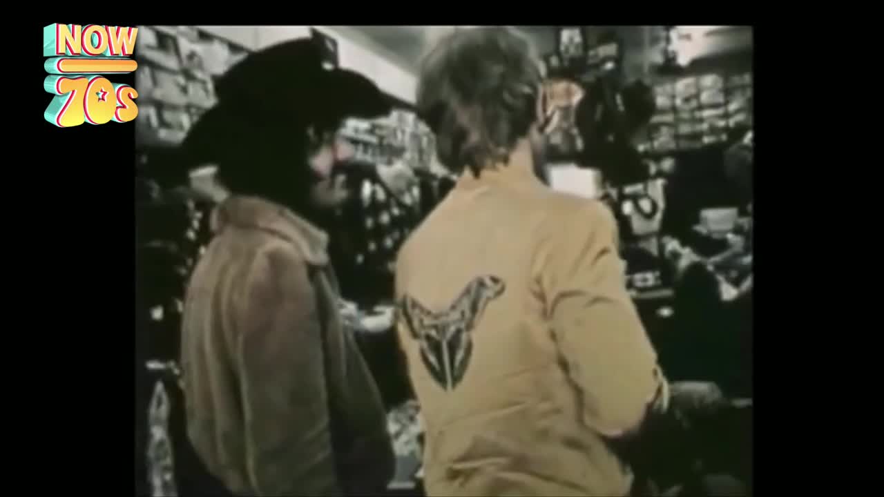 Two men, one in a tan jacket with a distinctive emblem on the back, are browsing in a shop. The shelves behind them are packed with items, and the lighting has a warm, vintage feel.
