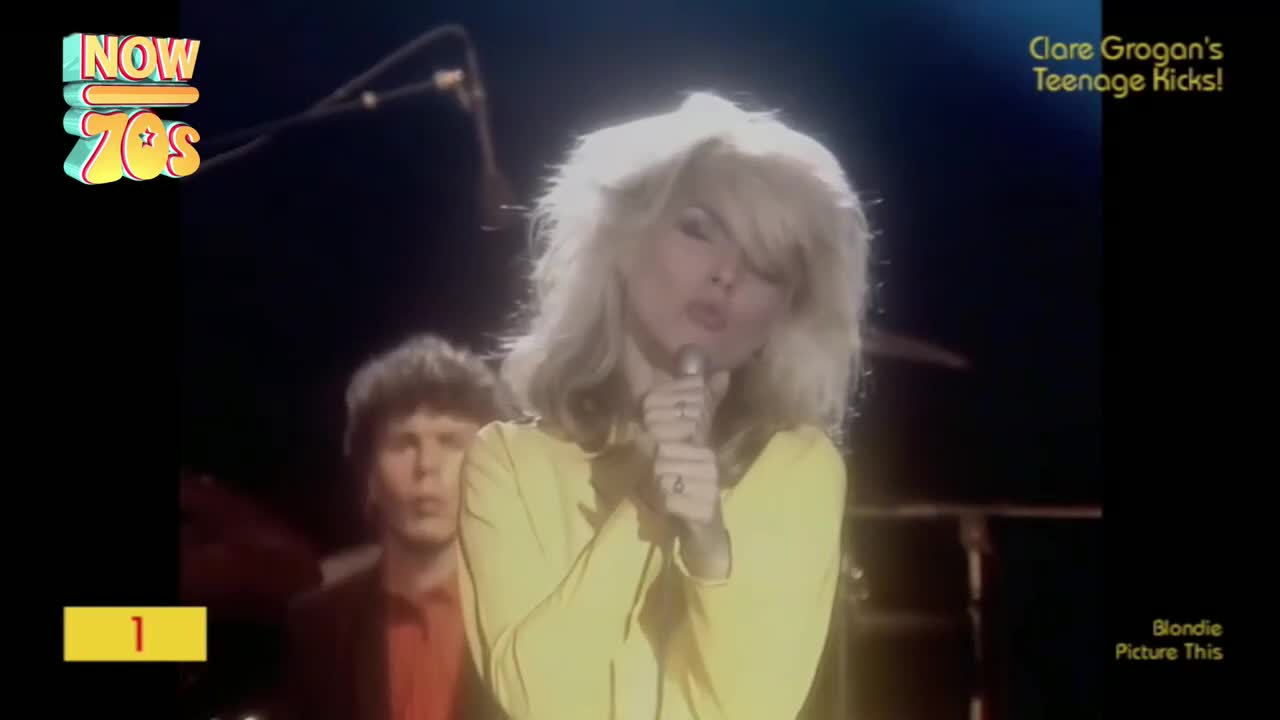 Blondie's frontwoman, in a bright yellow top, is singing into a microphone. Behind her, a band member in a red shirt looks on.

