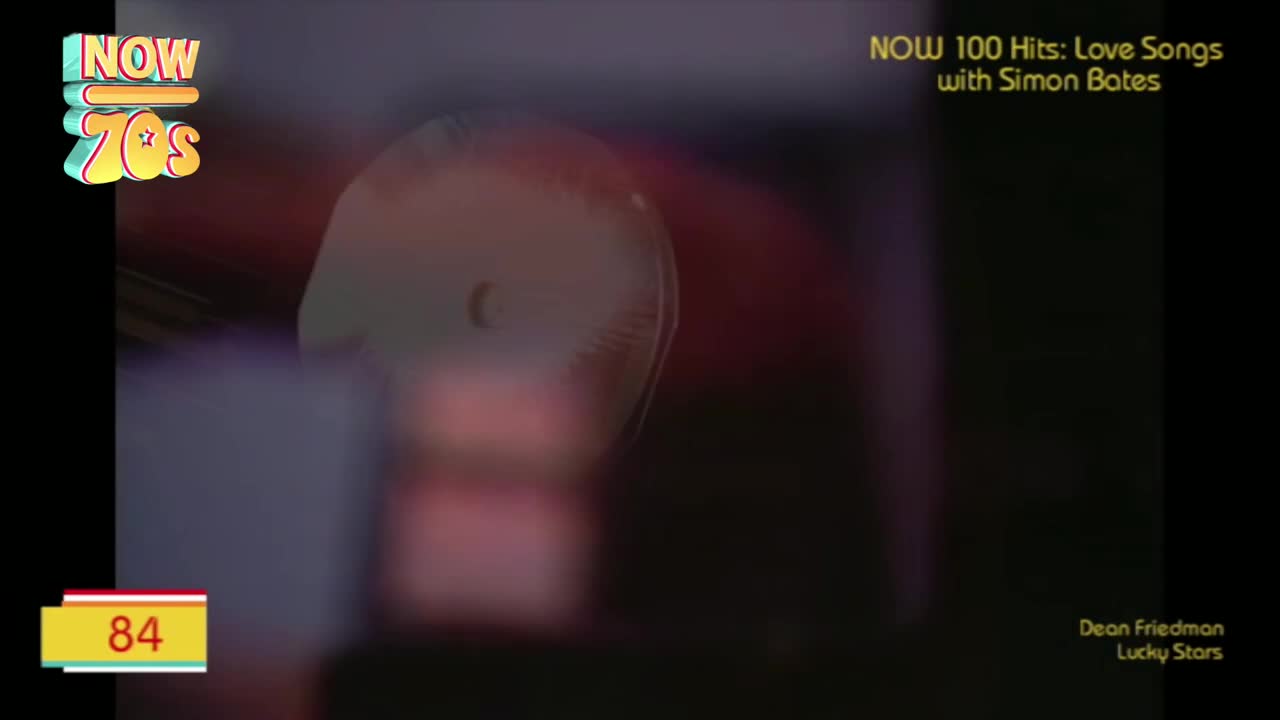 The "Now 70s" logo sits in the corner, while a blurred image of a fan spins in the background. The number 84 flashes on the screen, and the song "Lucky Stars" by Dean Friedman is playing.
