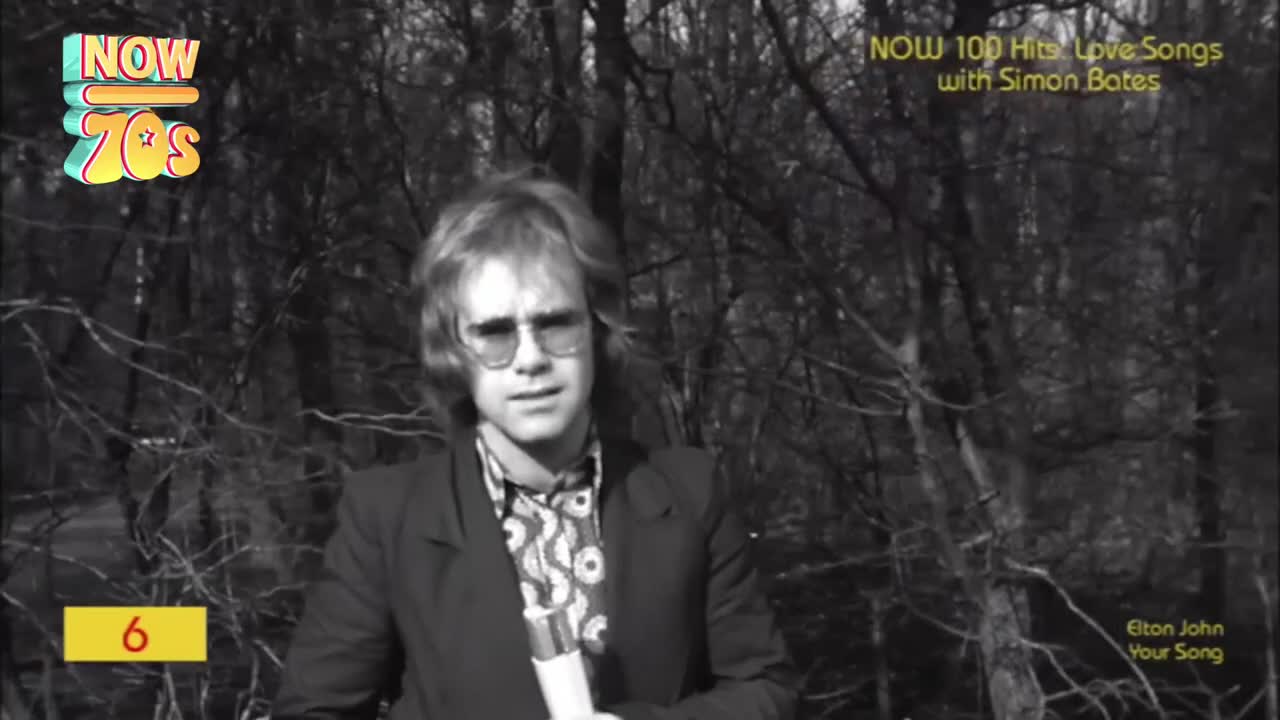 Elton John, in a patterned shirt and dark jacket, holds a microphone, standing against a backdrop of bare trees. The "Now 70s" logo and song title are visible, setting the scene for a music video.
