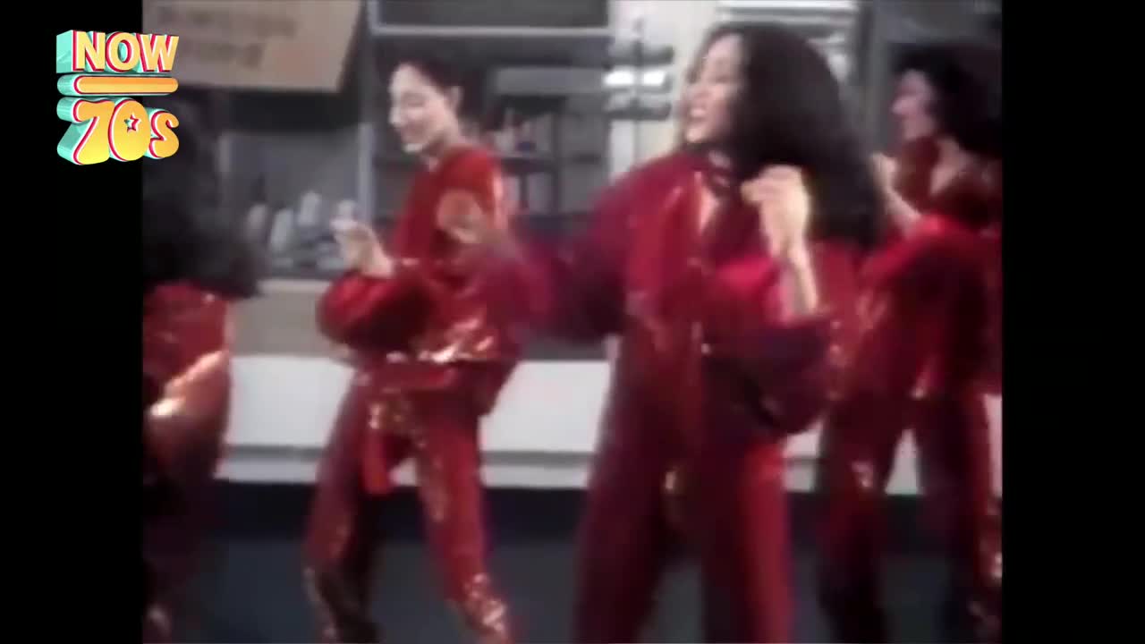 Women in shiny red jumpsuits are dancing, their arms swinging and bodies moving to the music. Their hair is flying, and they're all grinning from ear to ear.
