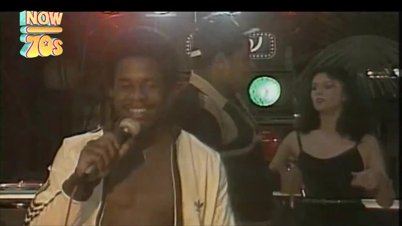 The singer, in a white Adidas jacket, holds a microphone, grinning as he belts out a tune. A woman in a black dress sways nearby, and the disco lights are flashing.
