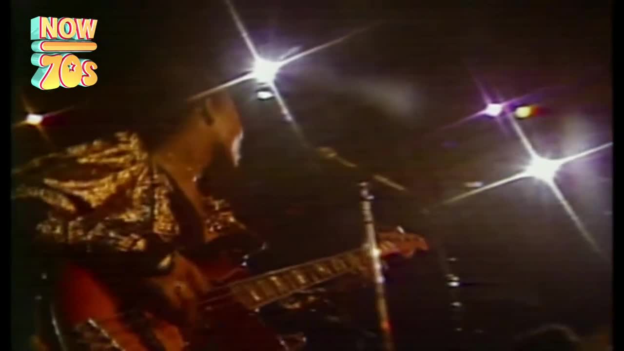 A musician in a shimmering gold jacket strums a bass guitar, bathed in the bright glare of stage lights. The microphone stand is close, and the music is loud.
