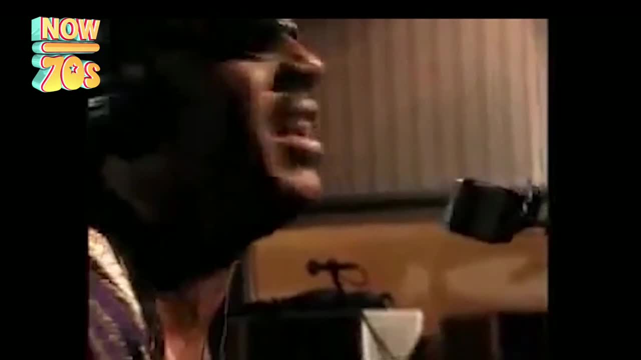 A man in a headband sings into a microphone, his mouth wide open. The studio lights catch the glint of sweat on his brow as he belts out a tune.
