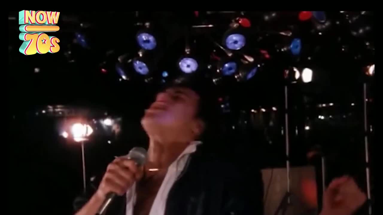 The singer holds a microphone, head thrown back, bathed in the glow of the stage lights. Overhead, the disco ball's reflections dance across the dark room.

