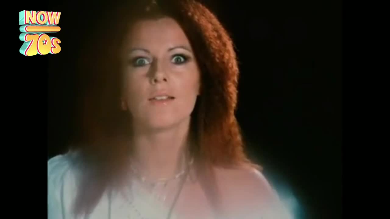 A woman with fiery red hair stares directly at the camera, her eyes wide with surprise. A soft, ethereal glow surrounds her as she appears to be in a darkened space.
