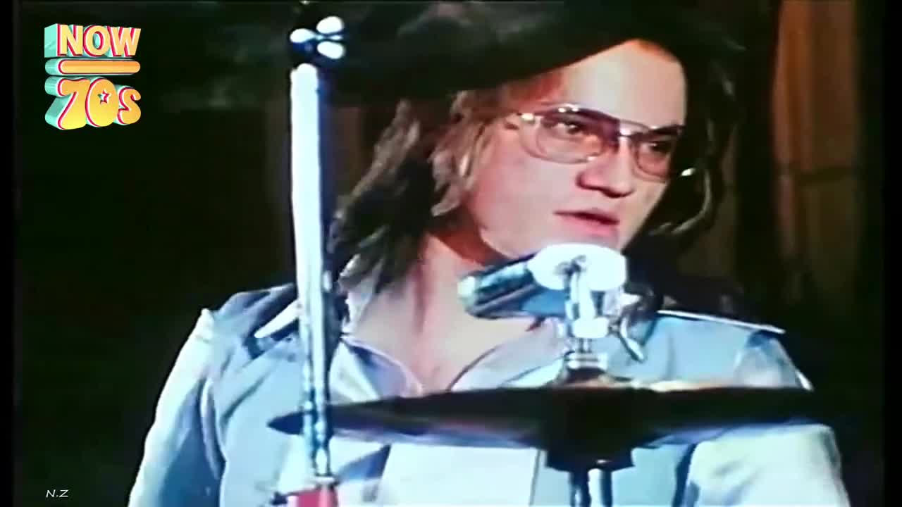 The drummer, sporting a beret and aviator glasses, is focused on the cymbals. His long hair is falling over his shoulders as he prepares to strike.
