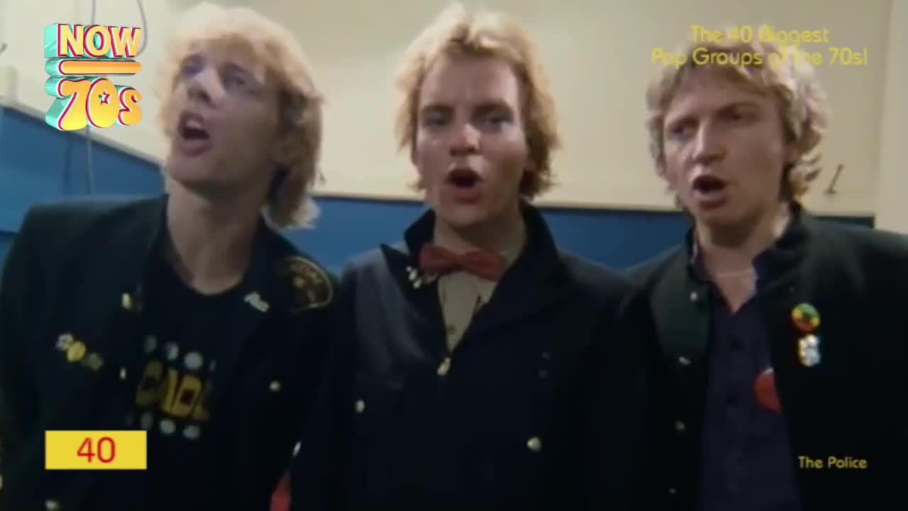 Three men with matching blond hair and dark jackets are singing, their mouths wide open. A small red bow tie adds a touch of flair to the center singer's outfit.
